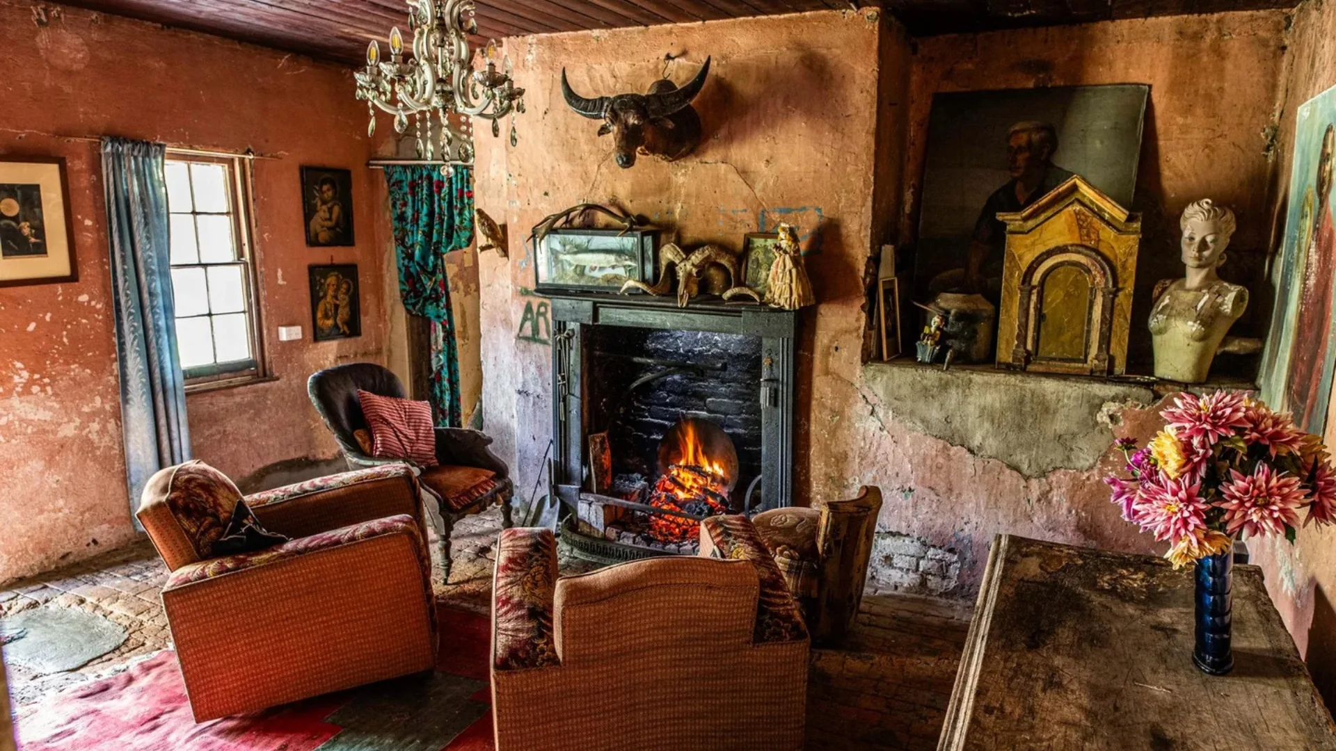 Cozy rustic room inside a Georgian house with a lit fireplace, antique decor, armchairs, and a vase of flowers on a table.