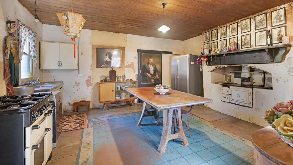 Kitchen inside a Georgian house with wooden table, blue rug, eclectic decor, hanging light, and framed portraits on distressed walls.