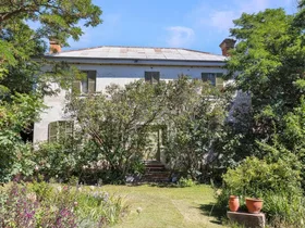 Large white house surrounded by lush green trees and plants, featuring a metal roof and shuttered windows.