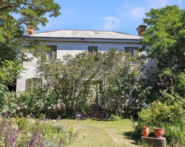 Large white house surrounded by lush green trees and plants, featuring a metal roof and shuttered windows.