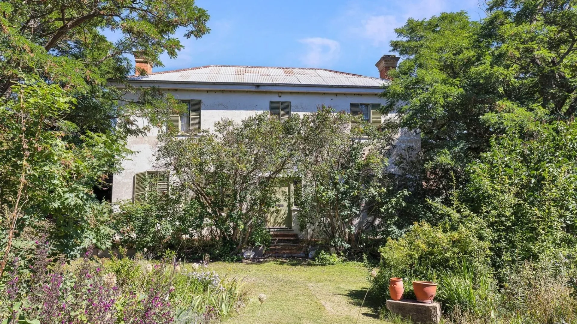 Large white house surrounded by lush green trees and plants, featuring a metal roof and shuttered windows.