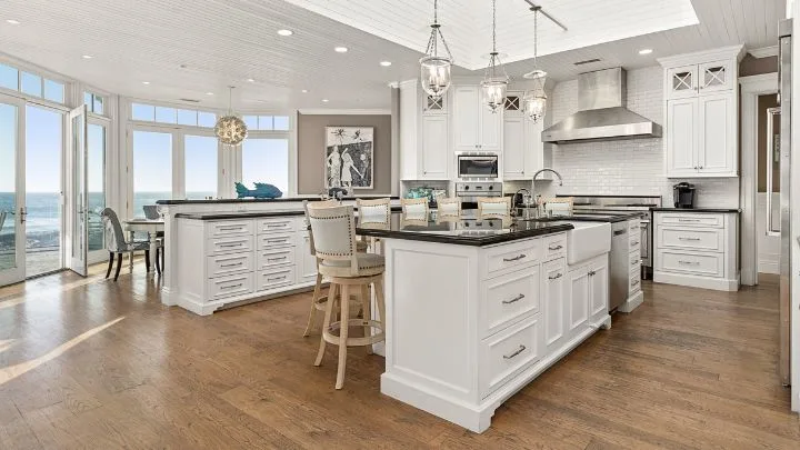 the kitchen in the real hannah montana house a sprawling kitchen space with two kitchen islands, black granite benchtops, white cabinets and timber floorboards with views of the ocean.