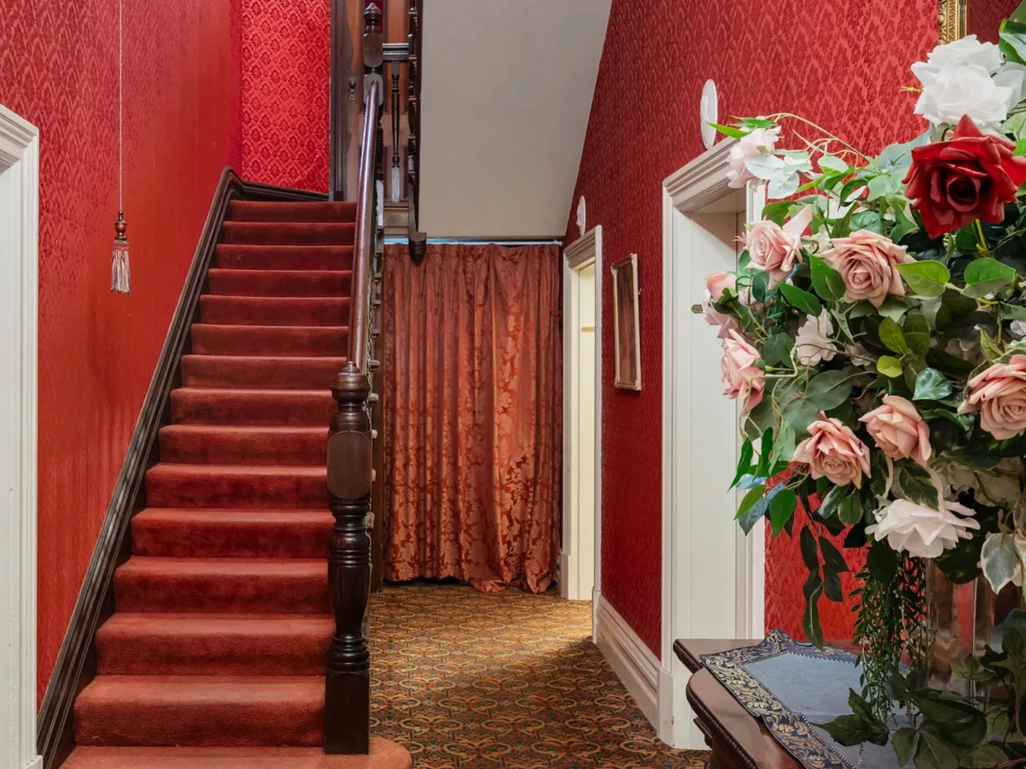 A red wall-papered interior with dark timber antique staircase and red carpet leading upstairs