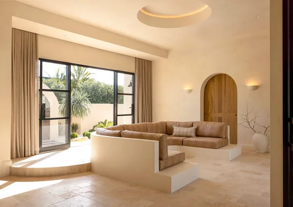 Beige living room with a large sectional sofa, arched wooden door, and glass doors opening to a garden view.