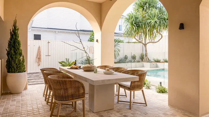 Stylish outdoor dining area with wicker chairs, stone table, and pool view under archways.