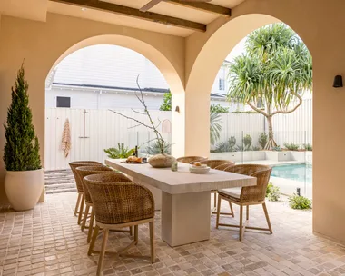 Stylish outdoor dining area with wicker chairs, stone table, and pool view under archways.