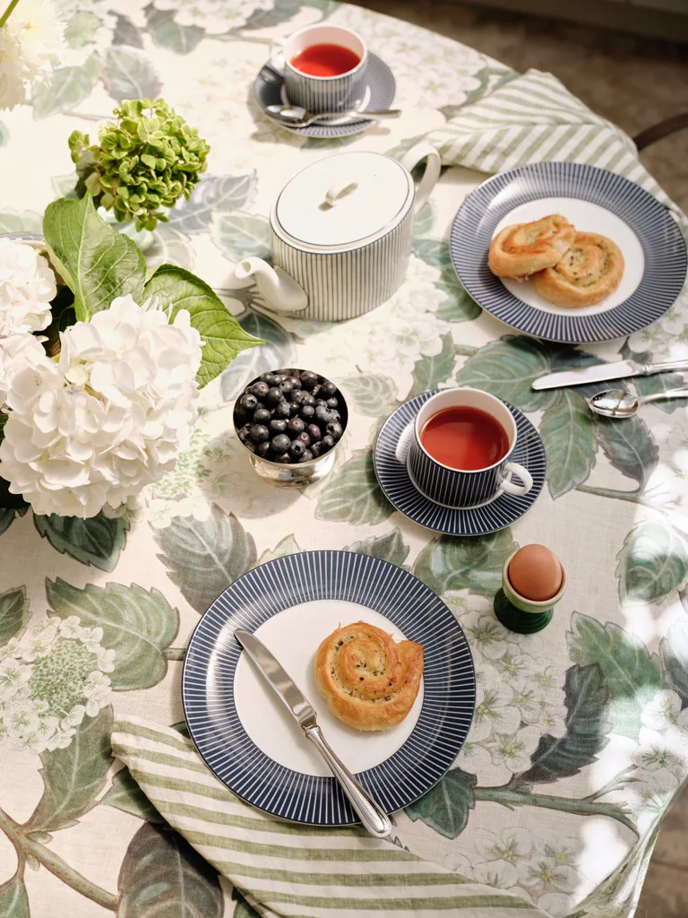 A printed floral tablecloth with green and white flowers, on a table set with blue and white plates and assortments for morning tea.