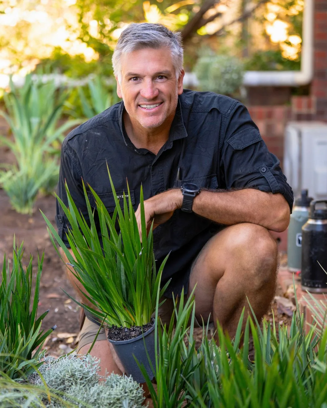 Dennis Scott from Selling Houses Australia poses crouched in the garden surrounded by green plants