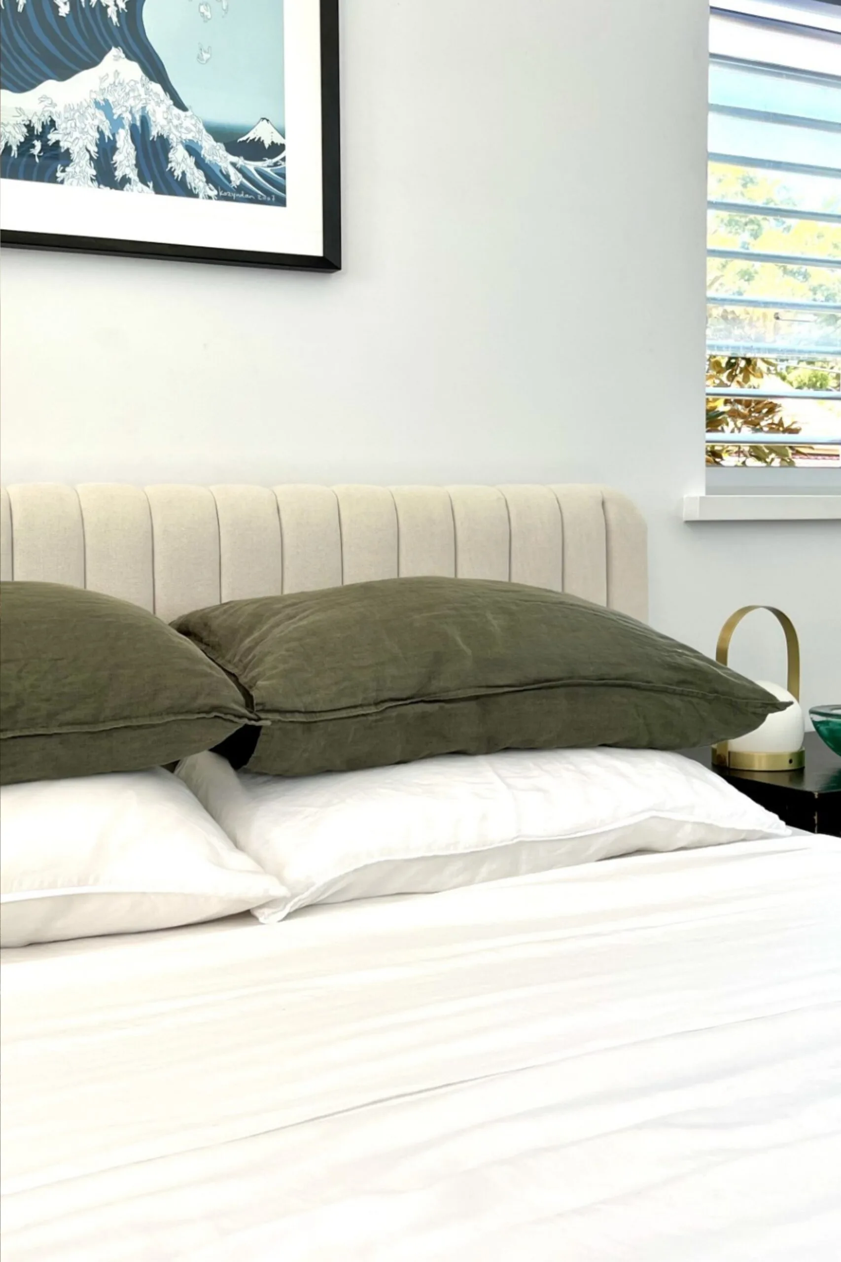 Bed with olive green linen pillows, white bamboo sheets, a beige bedhead and a framed wave art on the wall.