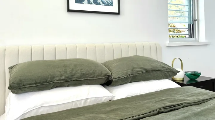 Beige bedhead with olive green linen pillows and bedding, white bamboo sheets and a framed wave artwork on the wall.