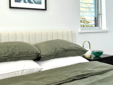 Beige bedhead with olive green linen pillows and bedding, white bamboo sheets and a framed wave artwork on the wall.