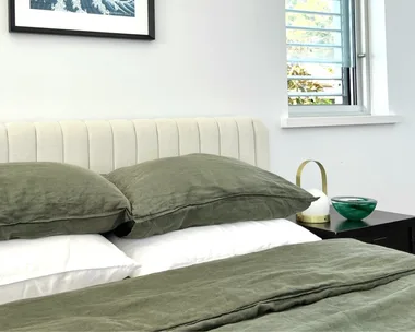 Beige bedhead with olive green linen pillows and bedding, white bamboo sheets and a framed wave artwork on the wall.