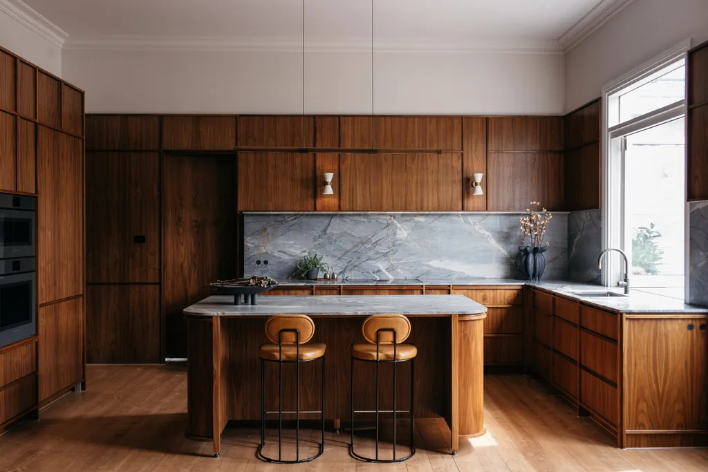 A mid-century style kitchen with timber cabinetry, grey toned marble benchtop and splashback. The same timber has been used on the profile of an island, with the same marble topping it. Two tan leather stools are positioned on one side of the island. A slim horizontal pendant light hands from the ceiling above the island, with two cone-shaped white wall sconces attached to timber panelling between upper cupboards.