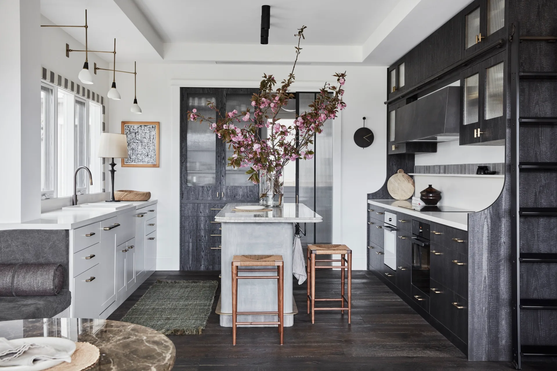 A custom and bespoke kitchen. Along one wall, white cabinetry is positioned underneath a white benchtop. The wall above has windows. The opposite wall features dark toned cabinetry with a white benchtop and splashback. The rangehood is also a dark tone. In the middle of the room is a grey toned island, small in size, with two timber stools positioned beside it. A vase with tall branches decorated with pink flowers is positioned on the island.