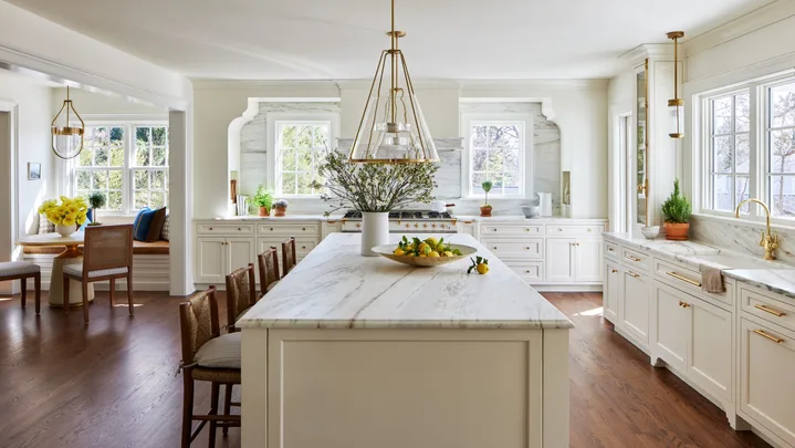 A large kitchen that features white on white finishes with brass details. White shaker-style joinery is paired with brass handles and knobs. Two brass pendant lights hang above a white panelled island. Every benchtop and splashback is comprised of the same white marble, with grey veining. In the left of the photo, a breakfast nook is positioned, with a built-in banquette window seat, two dining chairs and a round dining table. The floors are a deep toned timber.