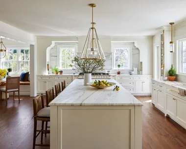 A large kitchen that features white on white finishes with brass details. White shaker-style joinery is paired with brass handles and knobs. Two brass pendant lights hang above a white panelled island. Every benchtop and splashback is comprised of the same white marble, with grey veining. In the left of the photo, a breakfast nook is positioned, with a built-in banquette window seat, two dining chairs and a round dining table. The floors are a deep toned timber.
