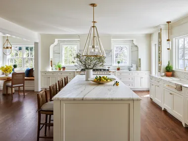 A large kitchen that features white on white finishes with brass details. White shaker-style joinery is paired with brass handles and knobs. Two brass pendant lights hang above a white panelled island. Every benchtop and splashback is comprised of the same white marble, with grey veining. In the left of the photo, a breakfast nook is positioned, with a built-in banquette window seat, two dining chairs and a round dining table. The floors are a deep toned timber.