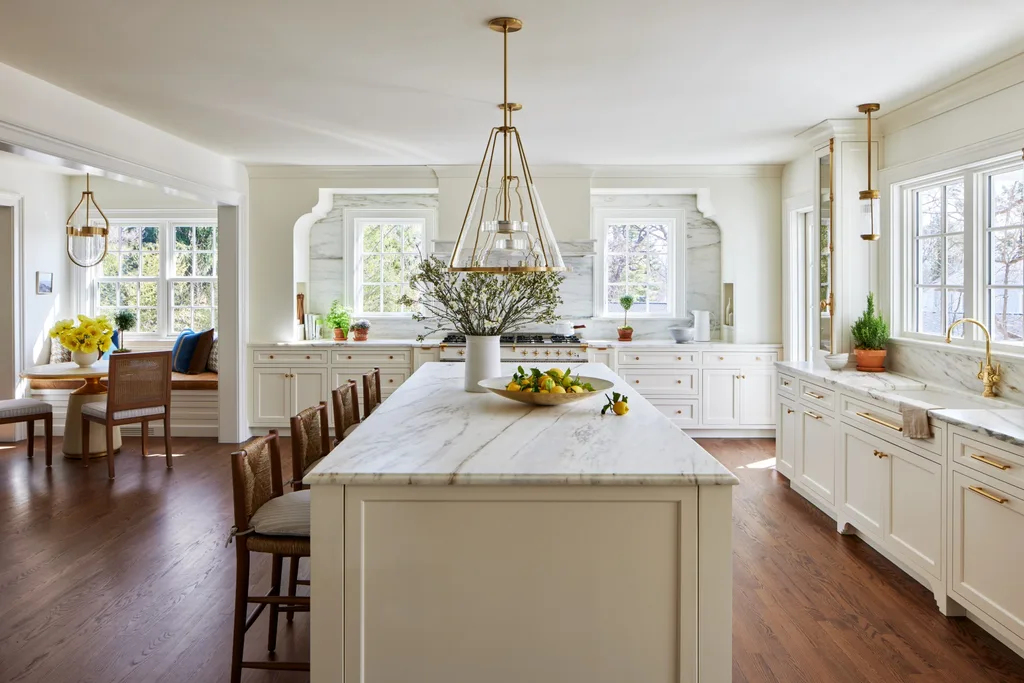 A large timeless kitchen that features white on white finishes with brass details. White shaker-style joinery is paired with brass handles and knobs. Two brass pendant lights hang above a white panelled island. Every benchtop and splashback is comprised of the same white marble, with grey veining. In the left of the photo, a breakfast nook is positioned, with a built-in banquette window seat, two dining chairs and a round dining table. The floors are a deep toned timber.