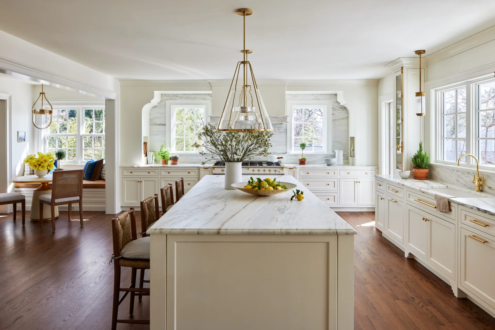 A large timeless kitchen that features white on white finishes with brass details. White shaker-style joinery is paired with brass handles and knobs. Two brass pendant lights hang above a white panelled island. Every benchtop and splashback is comprised of the same white marble, with grey veining. In the left of the photo, a breakfast nook is positioned, with a built-in banquette window seat, two dining chairs and a round dining table. The floors are a deep toned timber.