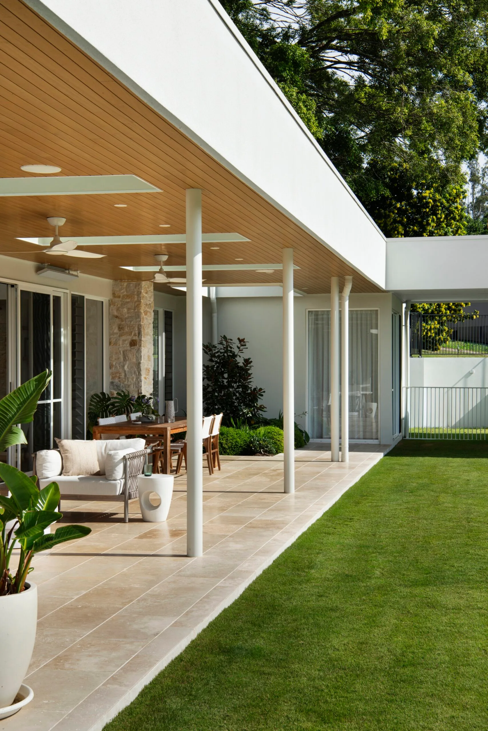 Modern patio with tiled floor, outdoor seating, ceiling fans, and lush plants, adjacent to a well-manicured lawn.