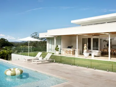 Modern house with glass doors, poolside loungers, umbrella, and inflatable ring in a clear sky setting.