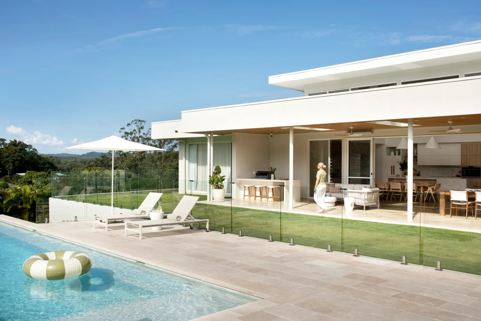 Modern house with glass doors, poolside loungers, umbrella, and inflatable ring in a clear sky setting.