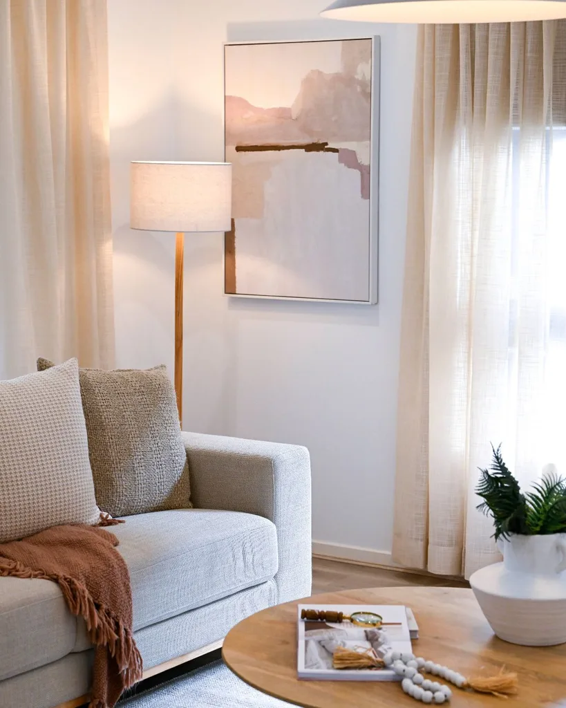 A living room scene with a lit standing lamp in the corner, lighting an abstract pink and beige artwork between windows with beige sheer curtains, and a grey lounge with a brown throw