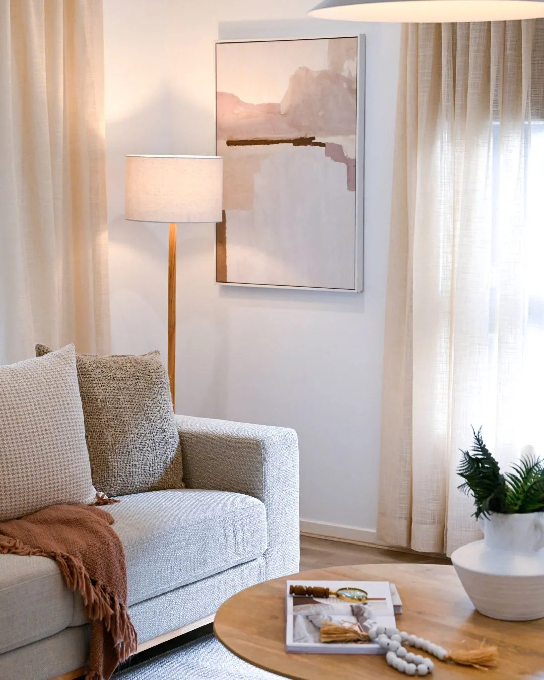 A living room scene with a lit standing lamp in the corner, lighting an abstract pink and beige artwork between windows with beige sheer curtains, and a grey lounge with a brown throw