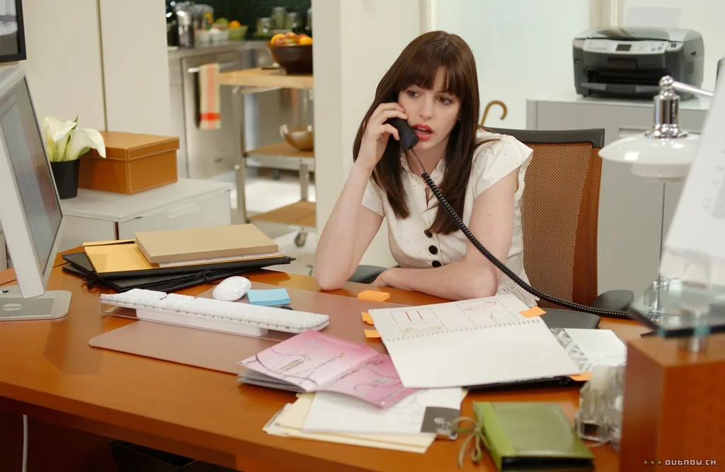 A still from the movie The Devil Wears Prada, featuring Anne Hathaway as Andy Sachs. The scene depicts Andy Sachs at her desk, which is a warm toned timber desk with a white keyboard and white desktop screen. The desk is cluttered with notebooks and Andy is on a black landline phone.