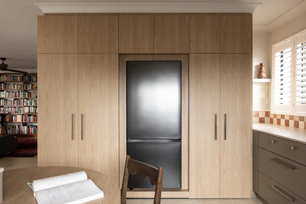 Built-in kitchen cabinet with integrated refrigerator, adjacent to bookshelves and a dining area with an open book. Underbench joinery in a taupe shade is to the right.