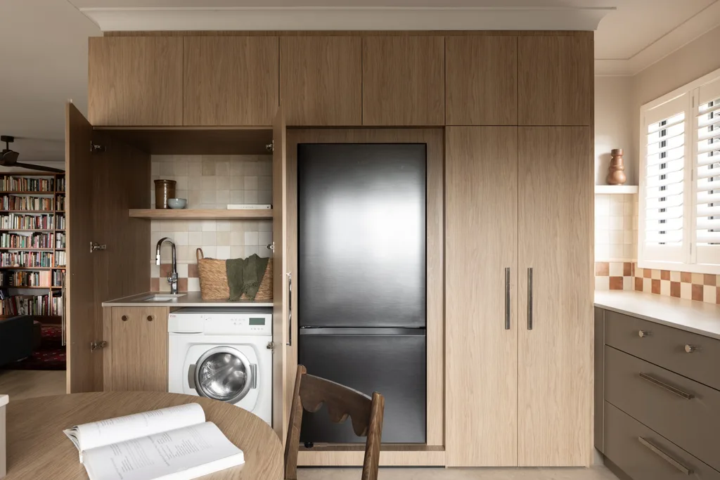 Built-in kitchen cabinets. Two cabinet doors are open to reveal integrated refrigerator, laundry nook including washing machine and sink, adjacent to bookshelves and a dining area with an open book. Underbench joinery in a taupe shade is to the right.
