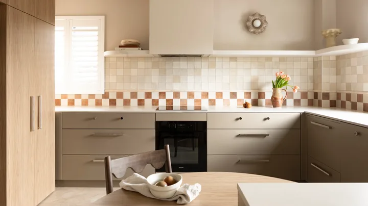 U-shaped kitchen with under-bench joinery in an earthy shade similar to taupe, tiled checkerboard backsplash, and vase of flowers on the counter. A small round wooden dining table is in the foreground, built into the corner of the cabinetry while tall cabinets are to the left of the image.