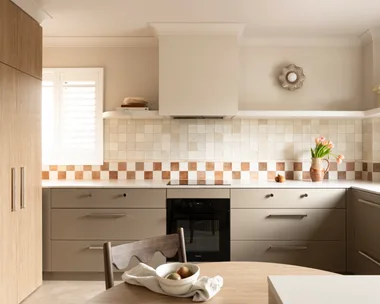 U-shaped kitchen with under-bench joinery in an earthy shade similar to taupe, tiled checkerboard backsplash, and vase of flowers on the counter. A small round wooden dining table is in the foreground, built into the corner of the cabinetry while tall cabinets are to the left of the image.