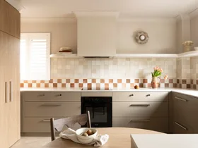 U-shaped kitchen with under-bench joinery in an earthy shade similar to taupe, tiled checkerboard backsplash, and vase of flowers on the counter. A small round wooden dining table is in the foreground, built into the corner of the cabinetry while tall cabinets are to the left of the image.
