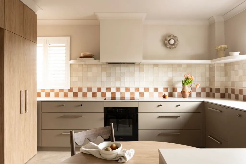 U-shaped kitchen with under-bench joinery in an earthy shade similar to taupe, tiled checkerboard backsplash, and vase of flowers on the counter. A small round wooden dining table is in the foreground, built into the corner of the cabinetry while tall cabinets are to the left of the image.
