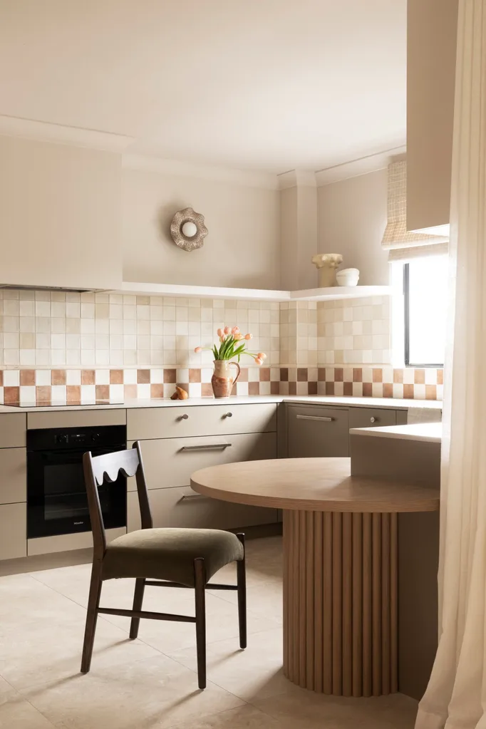 U-shaped kitchen with under-bench joinery in an earthy shade similar to taupe, tiled checkerboard backsplash, and vase of flowers on the counter. A small round wooden dining table is in the foreground, built into the corner of the cabinetry, with a dark dining chair. A vase with tulips is in the background on a benchtop.