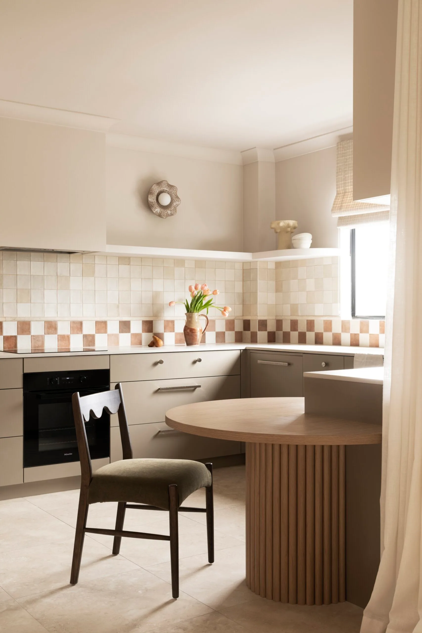 U-shaped kitchen with under-bench joinery in an earthy shade similar to taupe, tiled checkerboard backsplash, and vase of flowers on the counter. A small round wooden dining table is in the foreground, built into the corner of the cabinetry, with a dark dining chair. A vase with tulips is in the background on a benchtop.