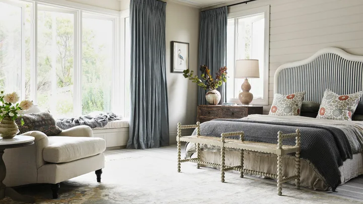 Bedroom with large windows, blue curtains, a cushioned armchair, striped headboard, and decorative pillows. A dark blue blanket on top of the bed matches the curtains, while a beige quilt is visible beneath. Two floral cushions are in front of dark green pillows.