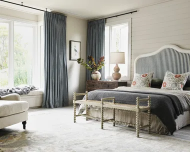 Bedroom with large windows, blue curtains, a cushioned armchair, striped headboard, and decorative pillows. A dark blue blanket on top of the bed matches the curtains, while a beige quilt is visible beneath. Two floral cushions are in front of dark green pillows.