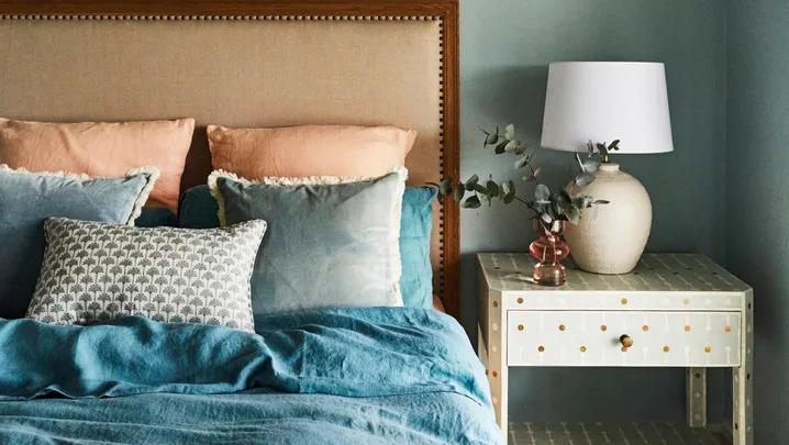 Bedroom with a cushioned headboard, blue bedding including a blue quilt cover, pillows, and a nightstand with a lamp and decorative vase.