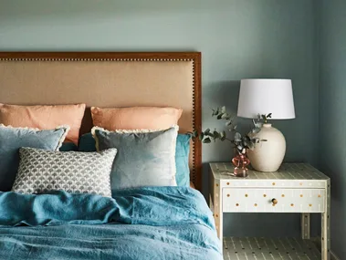 Bedroom with a cushioned headboard, blue bedding including a blue quilt cover, pillows, and a nightstand with a lamp and decorative vase.
