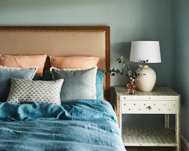 Bedroom with a cushioned headboard, blue bedding including a blue quilt cover, pillows, and a nightstand with a lamp and decorative vase.