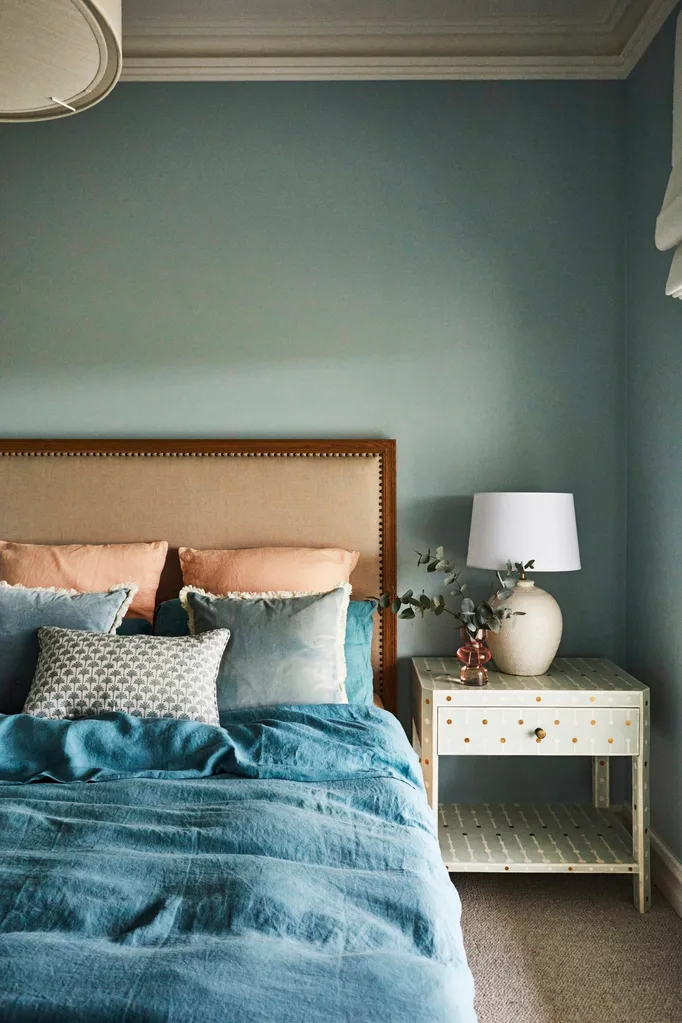 Bedroom with a cushioned headboard, blue bedding including a blue quilt cover, pillows, and a nightstand with a lamp and decorative vase.