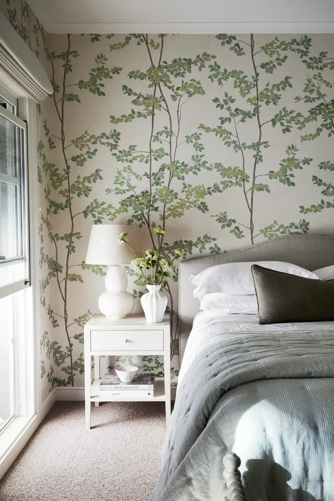 Bedroom with botanical-patterned wallpaper, a white bedside table, white lamp base and shade, white vase with flowers, and neatly made bed with cushions. The bed has a grey headboard and white pillows, with white bed sheets and blue quilt on top.