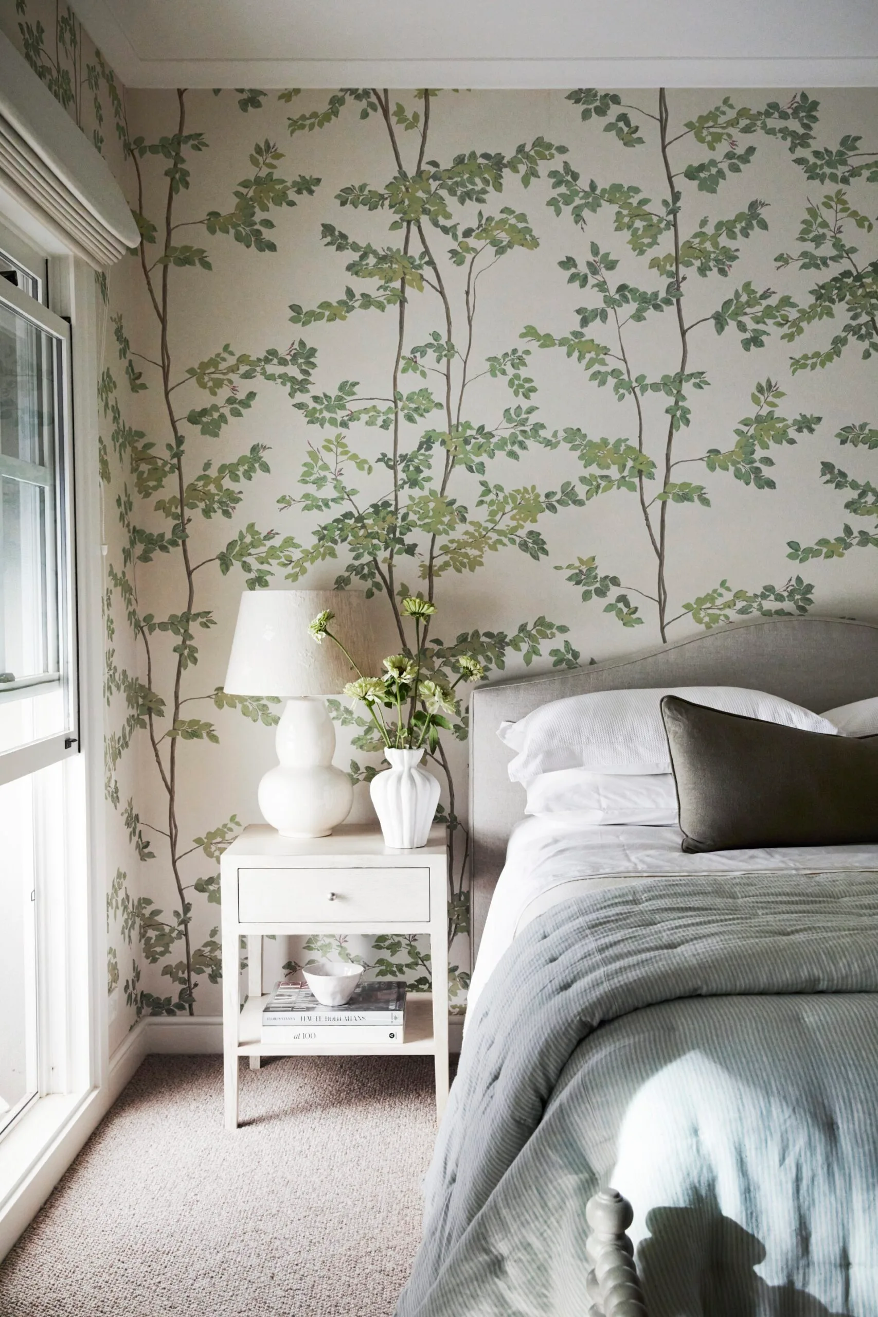 Bedroom with botanical-patterned wallpaper, a white bedside table, white lamp base and shade, white vase with flowers, and neatly made bed with cushions. The bed has a grey headboard and white pillows, with white bed sheets and blue quilt on top.