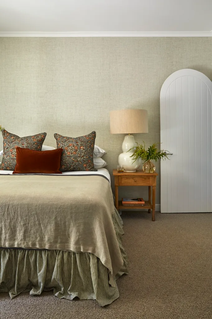 Cosy bedroom with a neatly made bed, floral decorative cushions, a timber bedside table with a lamp, and a white arched door. The bed has white pillows behind the floral cushions, a soft tan coloured black and white bedding underneath.
