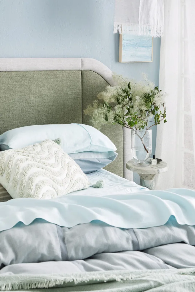 Bedroom with green headboard, blue bedding, decorative white pillow with a textured wavy pattern, plant in vase on marble nightstand, and pastel wall art. Bed sheets are light blue and the quilt on top is a darker shade of blue.