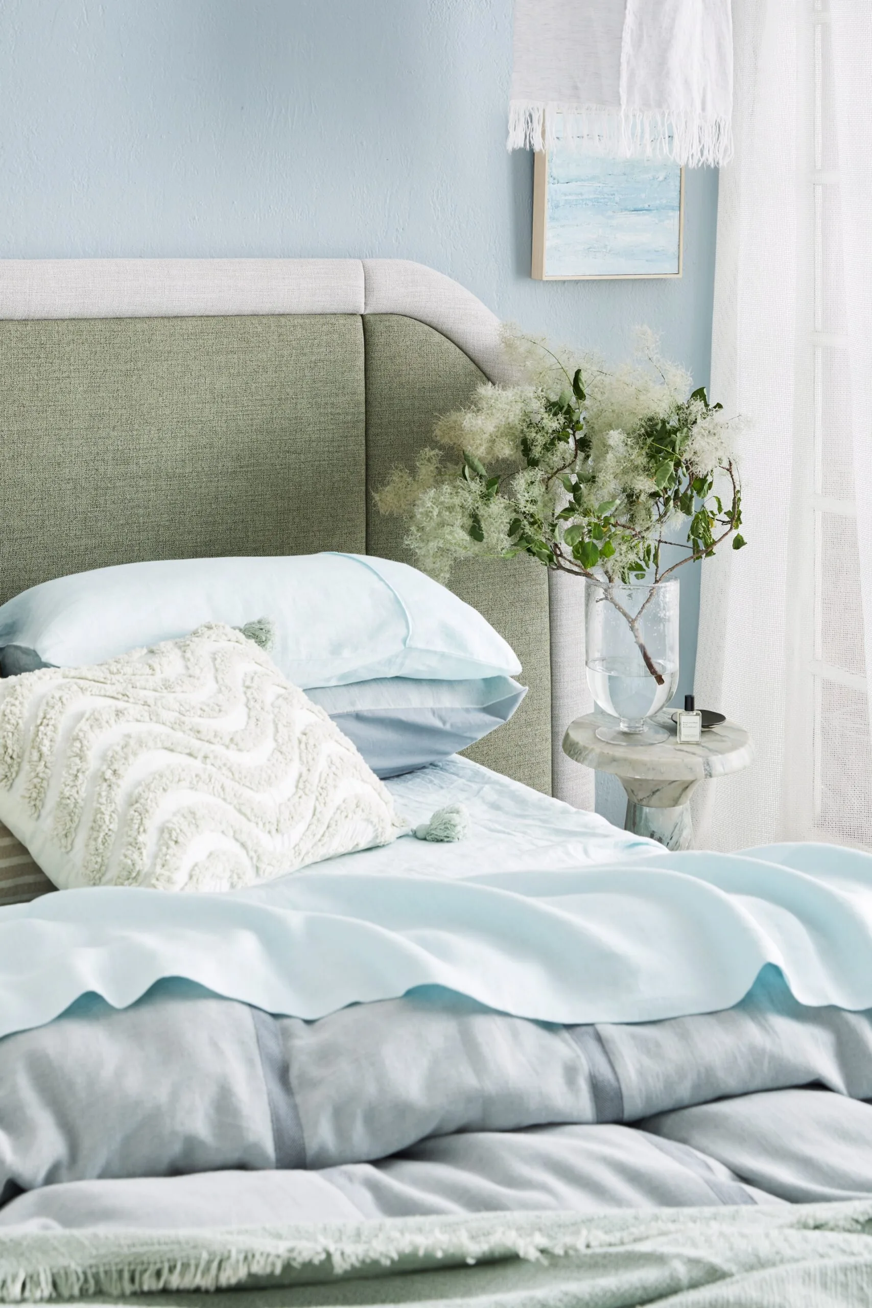Bedroom with green headboard, blue bedding, decorative white pillow with a textured wavy pattern, plant in vase on marble nightstand, and pastel wall art. Bed sheets are light blue and the quilt on top is a darker shade of blue.
