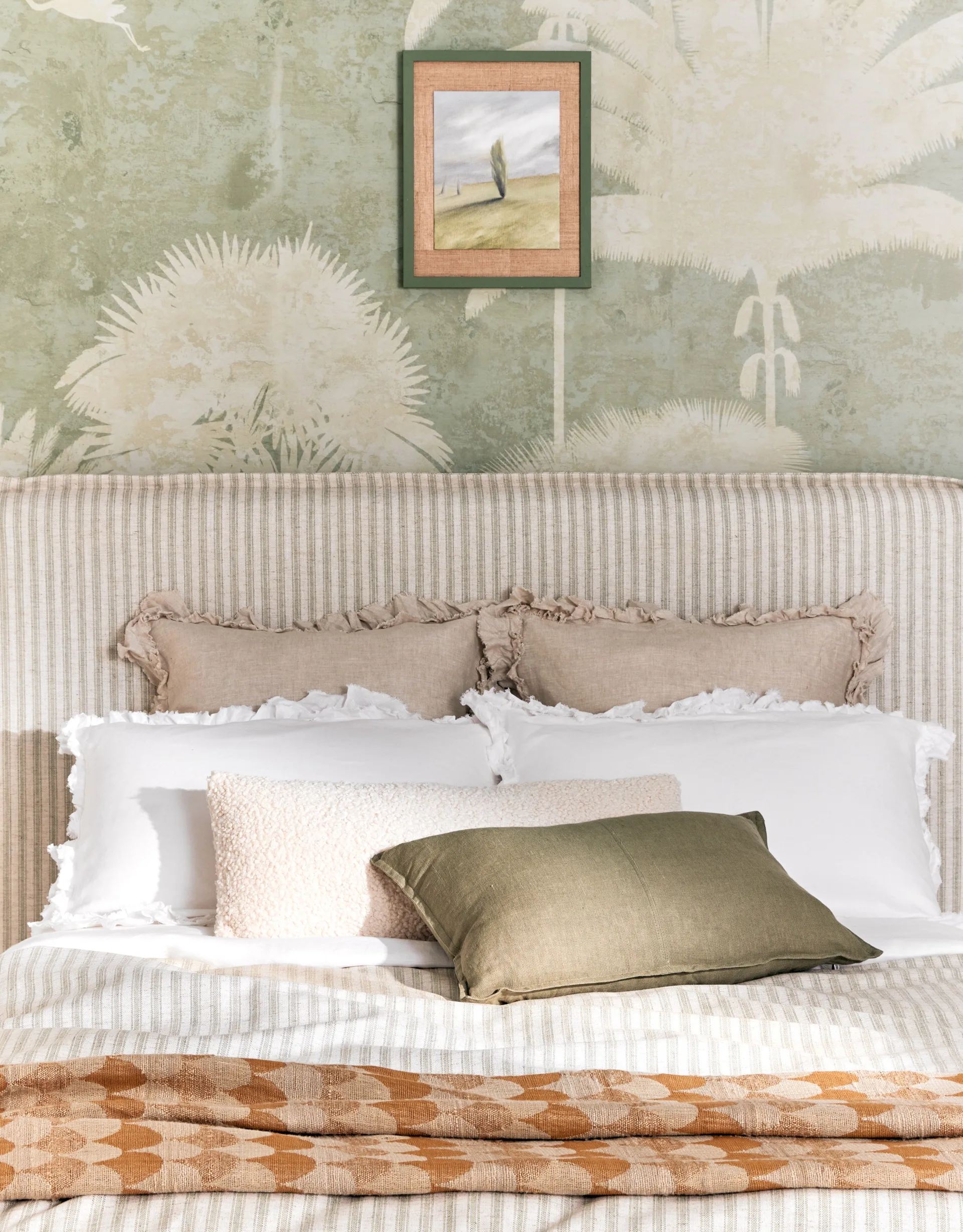Cozy bedroom with striped bedding, green pillow, and neutral cushions and pillows against a nature-themed wallpaper and framed art.