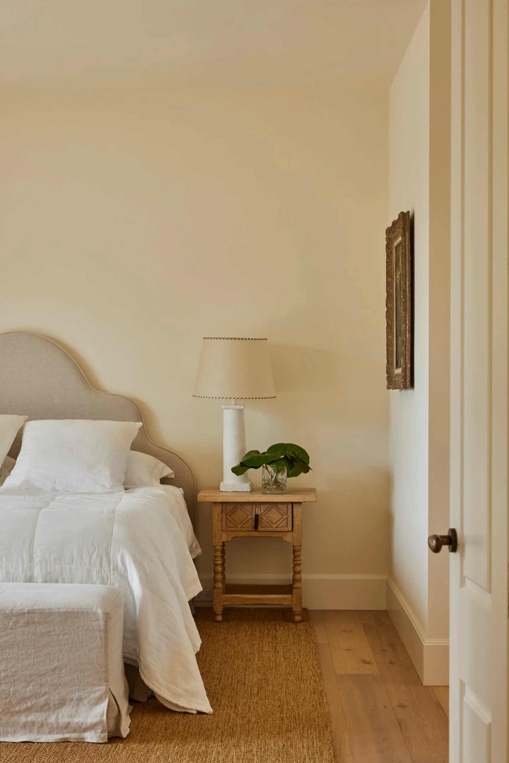 Cosy bedroom with warm white walls, white bedding including white quilt and white pillows, wooden nightstand, lamp, and plant decor on a woven rug.